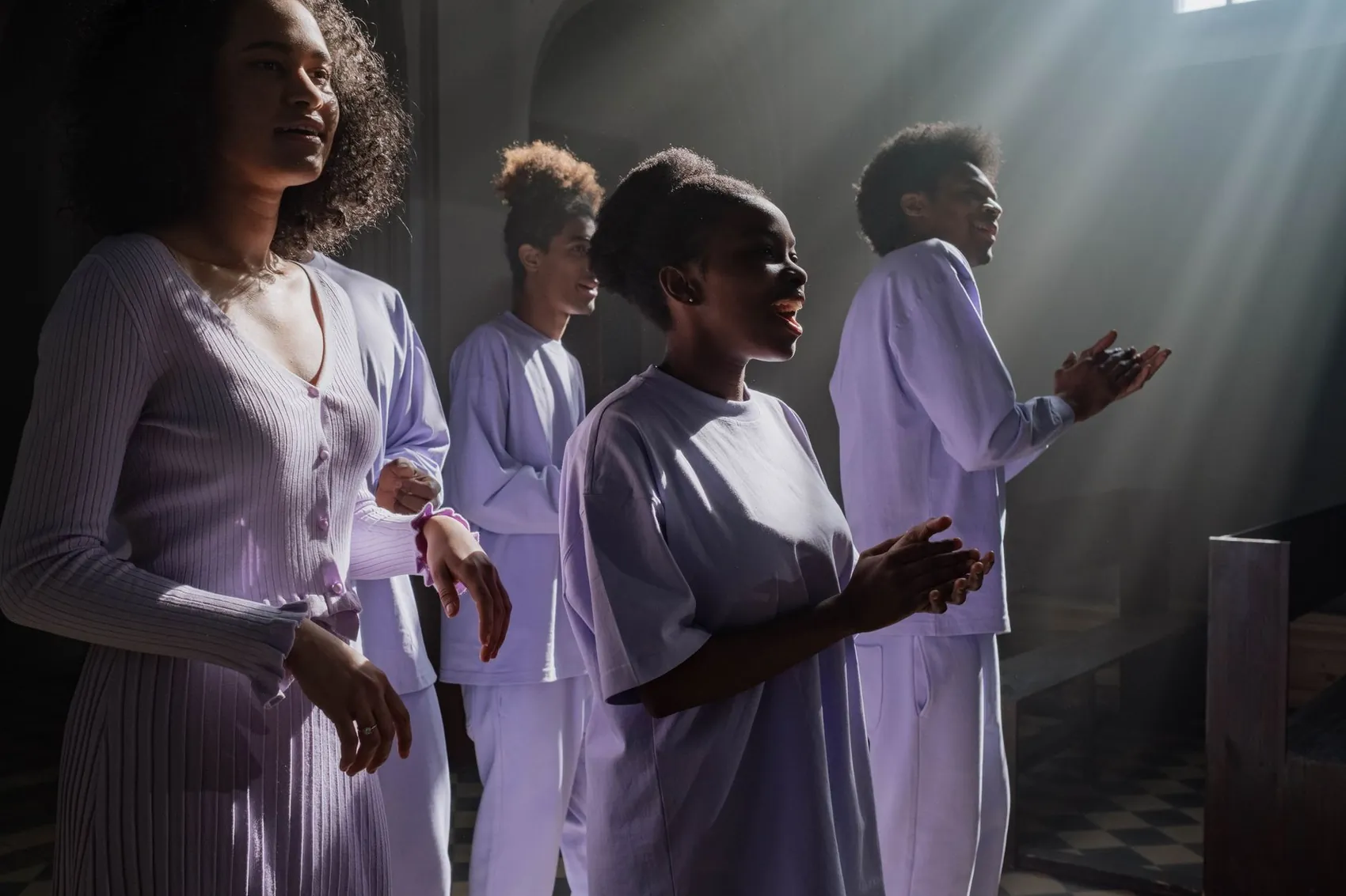 Four singers in lilac clothes clapping and singing in a church lit by beams of light