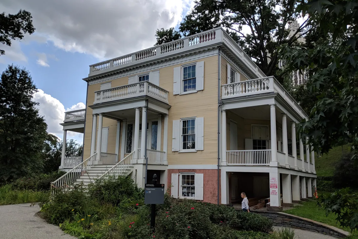 A pale yellow wooden house with white two-storey porches and balconies, set among trees on a curving path