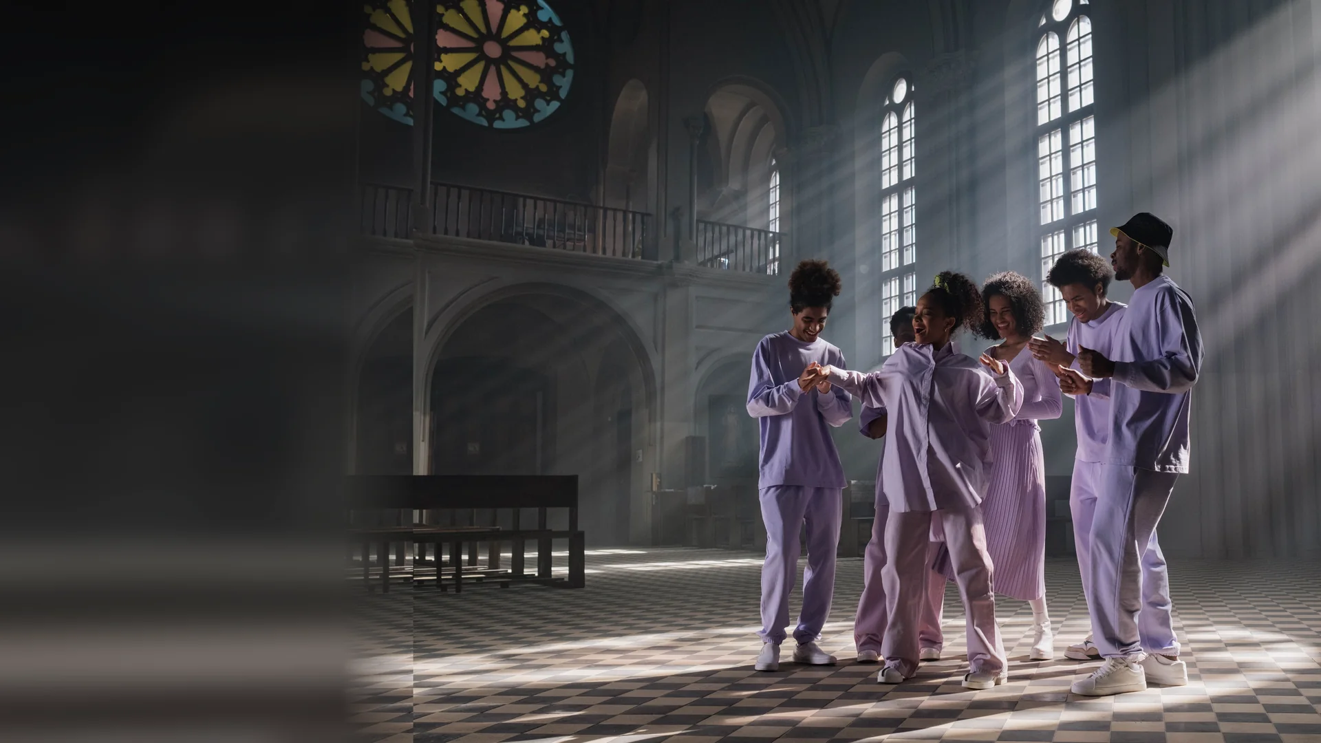 Singers in lilac clothes clapping and dancing together in a church hall, with shafts of sunlight falling through tall arched windows